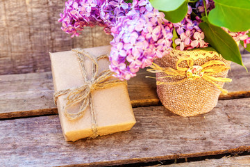 Fototapeta premium Floral decor elements. Small gift box with presents wrapped craft brown paper with bouquet of flowers beautiful smell violet purple lilac in vase on rustic wooden background. Copy space