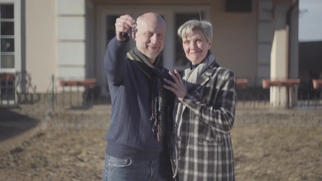 Happy senior couple, bald man and woman with grey short hair in coat and scarf stand near house and male showing a key in the hand. Bought new house. Family relationships.