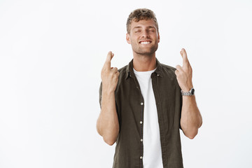 Excited optimistic and faithful good-looking sexy guy with fair hair and white smile grinning delighted with closed eyes as making wish with crossed fingers for good luck, hopefully praying