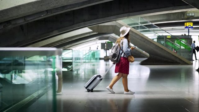 Young female passenger with suitcase and smartphone looking around while walking inside modern railway station