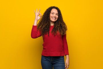 Teenager girl with red sweater over yellow wall showing ok sign with fingers