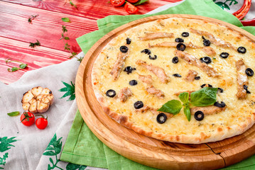 cheese pizza with chicken and olives on a round wooden board on a red wooden background, decorated with napkins, chili pepper and cherry tomatoes. close-up
