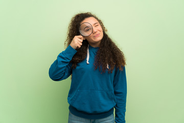 Teenager girl over green wall taking a magnifying glass and looking through it