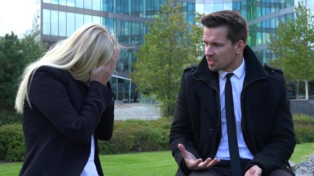 A businessman and a businesswoman sit on a bench and argue in an urban area - an office building in the background