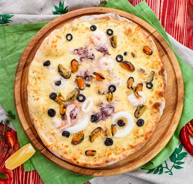 seafood pizza with shrimps, mussels, squids, olives and cheese on a round wooden board on a red wooden background, decorated with napkins, chili pepper and cherry tomatoes. close-up. top view