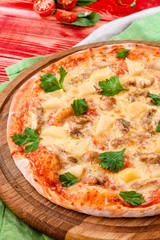 Pizza with chicken and pineapple on a round wooden board on a red wooden background, decorated with napkins, chili pepper and cherry tomatoes. close-up