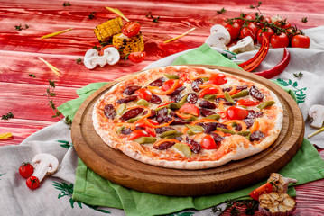 Italian Pizza with hunting sausages, tomatoes, chili peppers, cheese and pickles on a round wooden board on a red wooden background, decorated with napkins, chili pepper and cherry tomatoes. close up