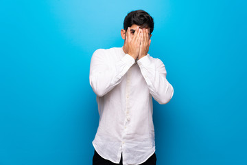 Young man over isolated blue wall covering eyes by hands and looking through the fingers