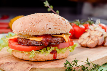 burger with juicy cutlet, tomatoes, cheese, lettuce and red sauce on a wooden board on a red wooden background, decorated with napkins, chilli pepper and cherry tomatoes. close up