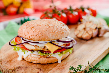 burger with chicken cutlet, tomatoes, cheese, lettuce and red sauce on a wooden board on a red wooden background, decorated with napkins, chili pepper and cherry tomatoes. close up