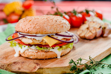 burger with chicken cutlet, tomatoes, cheese, lettuce and red sauce on a wooden board on a red wooden background, decorated with napkins, chili pepper and cherry tomatoes. close up