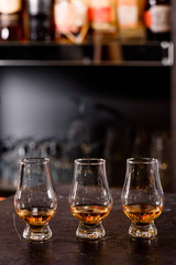Three Glencairn Glass with whiskey on a bar wooden counter close up on the background of blurry bottles. Close up. Dark background