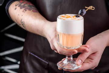 barman in a brown leather apron with a tattoo on his hand holding a glass with cocktail with white foam, decorated with a small plum. close up. space