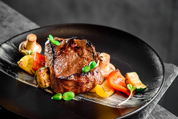 Juicy steak Filet Mignon with grilled vegetables served on a black plate. Mushrooms, zucchini, and sweet peppers are grilled. Close-up. Dark background. Space. Photo for the menu