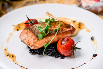 Beautiful serving of roasted salmon fillet and risotto with cuttlefish ink on a white plate with a signature sauce and Cherry tomatoes in a restaurant. close up