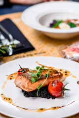 Beautiful serving of roasted salmon fillet and risotto with cuttlefish ink on a white plate with a signature sauce and Cherry tomatoes in a restaurant. close up