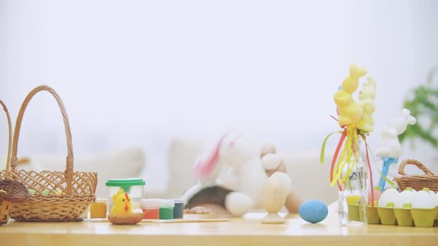 Little boy is hiding under the wooden table, full of Easter decorations: basket, yellow chicken, colorful eggs pains and paint-brush. Boy is playing with a cute, soft white bunny with pink ears, on