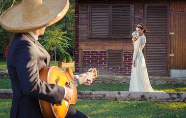 Groom with sombrero hat singing to his bride to be, playing guitar. Bride in the background holding...