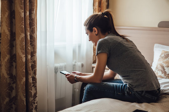 The girl sits on a bed in a hotel room and looks into the phone.