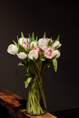 A bouquet of tulips of different colors, white, pink, stands in a glass vase in a dark interior.