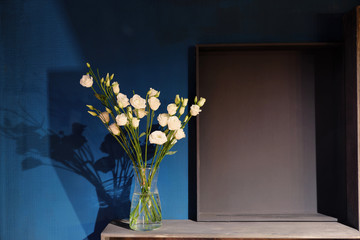 A bouquet of white roses in a glass vase stands against a dark blue wall. Wooden shelves.