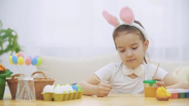 Little cute girl is having fun holding a paint-brush in her left hand. Girl with a beauty spots at her face watching at nude paint-brushes, sitting at the wooden table with Easter decorations. Girl is