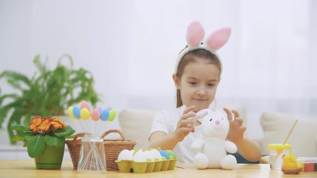 Brisk, cute girl is having fun painting with Easter bunny, which has the same ears, as she has. Girl is setting back white bunny. Girl with a beauty spot at her face and is smiling widely, sitting at