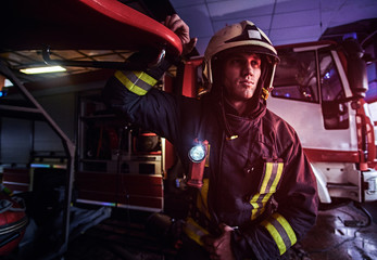 Portrait of a handsome fireman wearing a protective uniform with flashlight included standing in a fire station garage and looking sideways