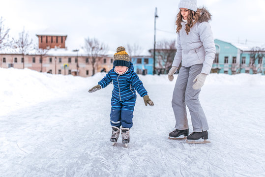 A young mother skates in park in winter, with her son a young child 3-5 years old. Holiday weekend at a public skating rink, snow and drifts in the background.