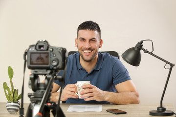 Young smiling cheerful man sitting at the office desk in front of the camera making a video. Male...