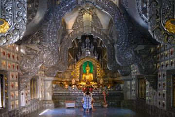 Interior view inside Thai temple of man sit, worship and pray in front of shrine and Golden Buddha statue.  Delicate Asian ornament and stainless steel decorated inner of Buddhist sanctuary. 