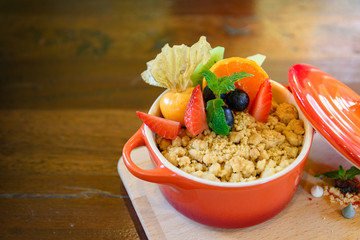 Apple Crumble in small ceramic orange mold pot and topping with fruit, strawberry, orange and blackberry, and cream on side, on wooden tray and table. 