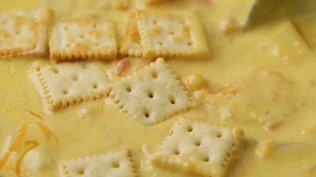 Close video of stirring crackers into chicken corn chowder with cheese then taking a spoonful at the end illuminated with natural lighting.