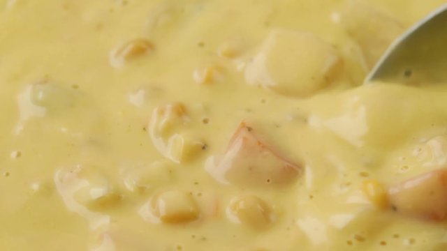 Close video of stirring hot chicken corn chowder in a pan while heating with a spoon and taking a spoonful at the end illuminated with natural lighting.