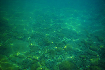 Fototapeta na wymiar Gravel bottom in shallow green sea with areas of larger rocks and boulders and reflections from surface.