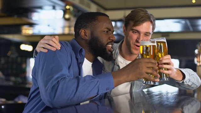 Two young men watching sport competition in pub, clinking beer glasses, hobby