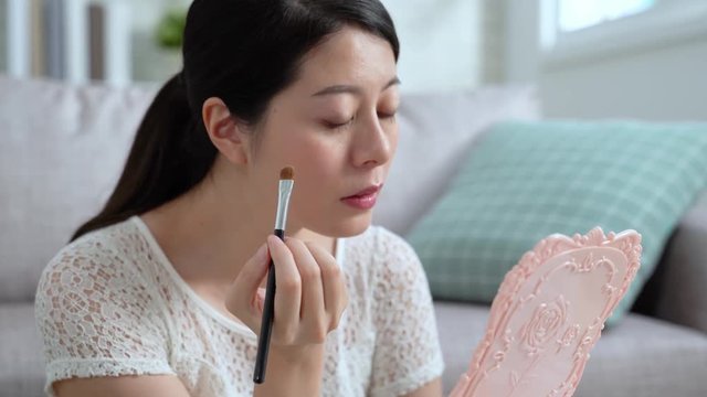 Attractive asian woman using brush and looking at mirror to makeup for night party sitting in home sofa in apartment living room. young girl blogger concentrated teach showing how to apply eyebrows