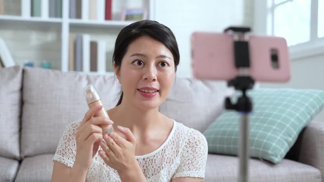 young asian female vlogger holding foundation introducing cosmetic recording online video for internet shop. beautiful girl showing makeup in live streaming by mobile phone talking to fans.
