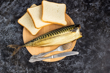 Smoked mackerel without head with fork knife cutting board bread on black background