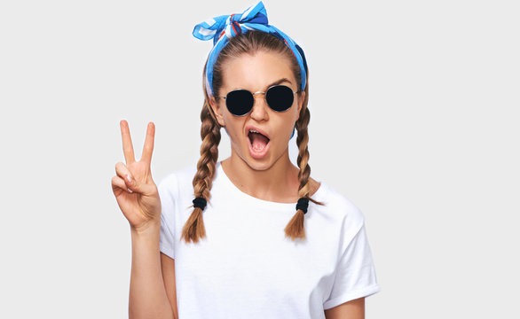 Horizontal studio portrait of cheerful blonde young woman wearing trendy sunglasses, white t-shirt and blue headband, showing peace sign. Student girl going funny with braids hairstyle. People emotion