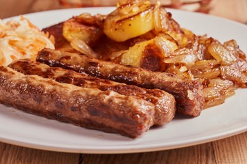 Serbian cevapi, cevapcici, Balkan minced meat kebab on a white plate with marinated cabbage, fried potatoes and fried onions on a wooden background. Close-up
