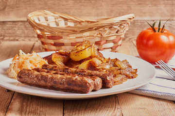 Serbian cevapi, cevapcici, Balkan minced meat kebab on a white plate with marinated cabbage, fried potatoes and fried onions next to bread, tomat and cutlery on a wooden background