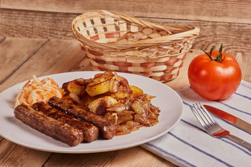Serbian cevapi, cevapcici, Balkan minced meat kebab on a white plate with marinated cabbage, fried potatoes and fried onions next to bread, tomat and cutlery on a wooden background