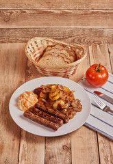 Serbian cevapi, cevapcici, Balkan minced meat kebab on a white plate with marinated cabbage, fried potatoes and fried onions next to bread, tomat and cutlery on a wooden background