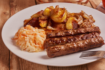 Serbian cevapi, cevapcici, Balkan minced meat kebab on a white plate with marinated cabbage, fried potatoes and fried onions next to fork on a wooden background