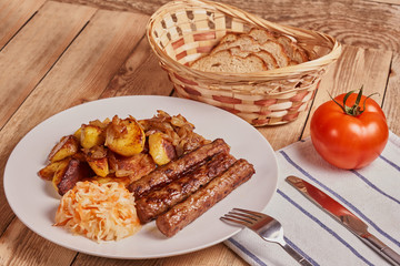 Serbian cevapi, cevapcici, Balkan minced meat kebab on a white plate with marinated cabbage, fried potatoes and fried onions next to bread, tomat and cutlery on a wooden background