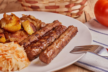 Serbian cevapi, cevapcici, Balkan minced meat kebab on a white plate with marinated cabbage, fried potatoes and fried onions next to bread, tomat and cutlery on a wooden background