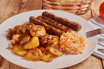 Serbian cevapi, cevapcici, Balkan minced meat kebab on a white plate with marinated cabbage, fried potatoes and fried onions next to bread, tomat and cutlery on a wooden background