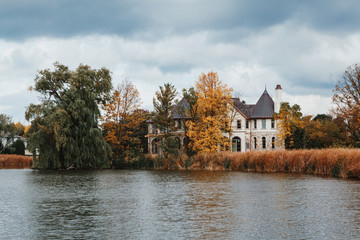 Fototapeta na wymiar Landscape nature view of beautiful Erupean gothic looking house castle hidden behind trees near pond lake with weeping willow trees and reed grass. Bad autumn weather with clouds in sky.