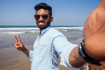 Young african american man in blue shirt and jeans making a selfie portrait on the front camera of a smartphone on the ocean beach.